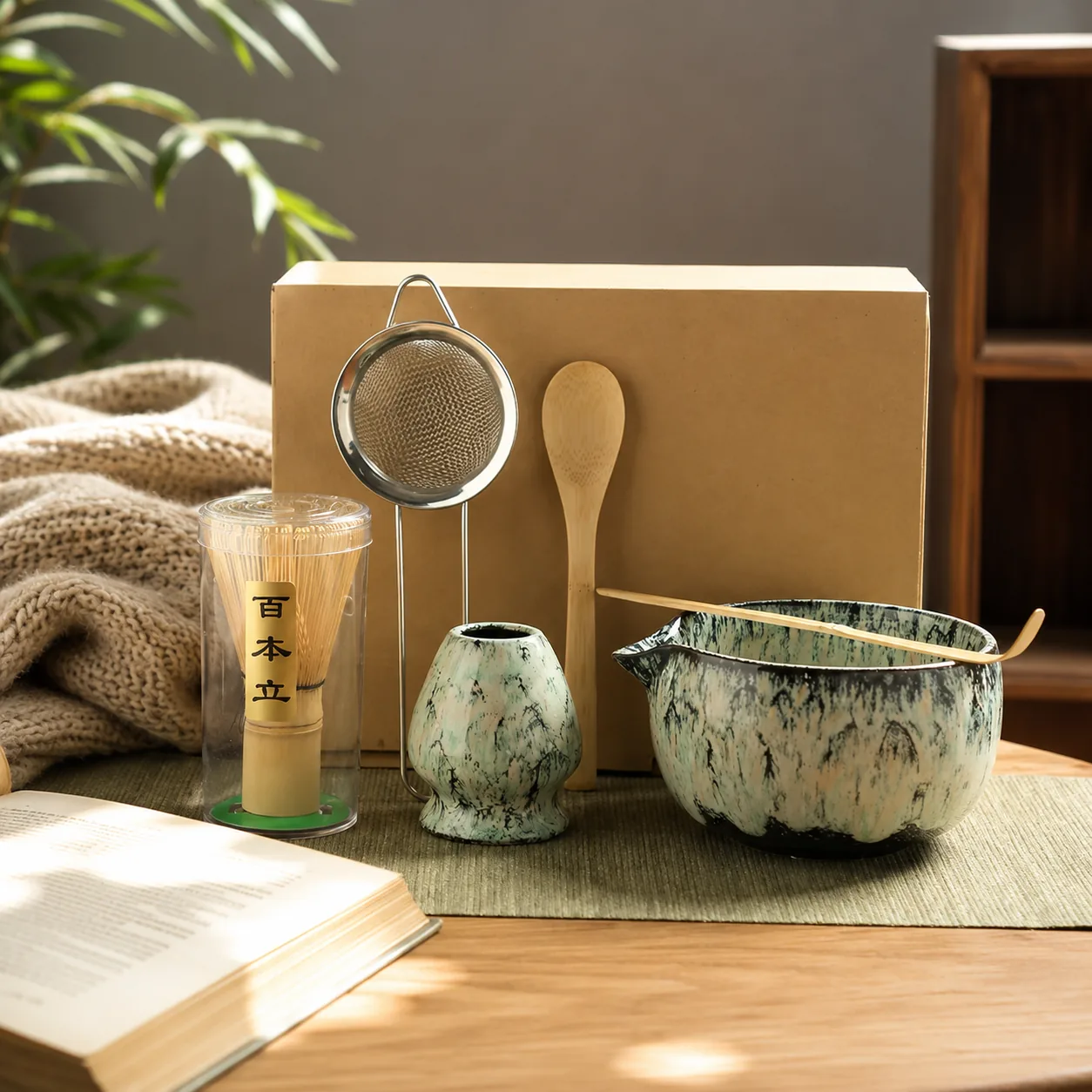 A green speckled matcha bowl with a bamboo scoop resting across it, next to a whisk in its case, a whisk holder and a sieve beside an open book in warm sunlight