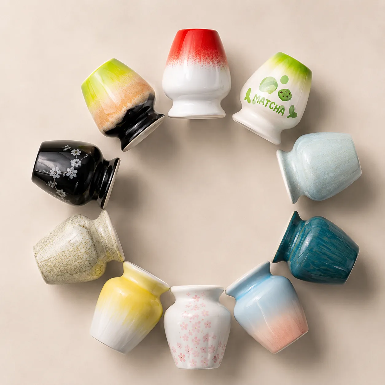 Ten ceramic matcha whisk holders in different glaze colors arranged in a ring and photographed from above on a cream background