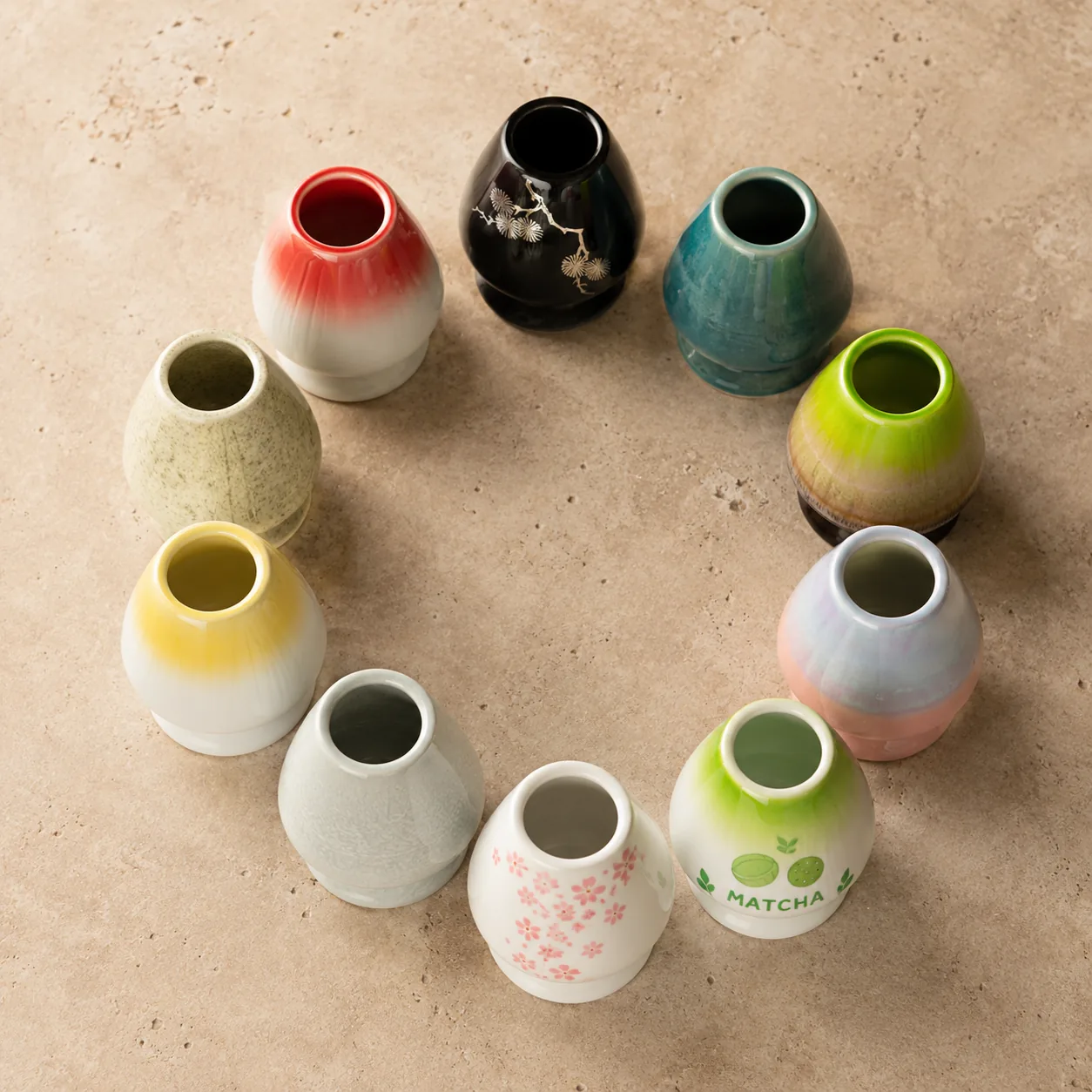 Ten ceramic matcha whisk holders in different glaze colors standing upright in a circle and photographed from above on a warm stone surface