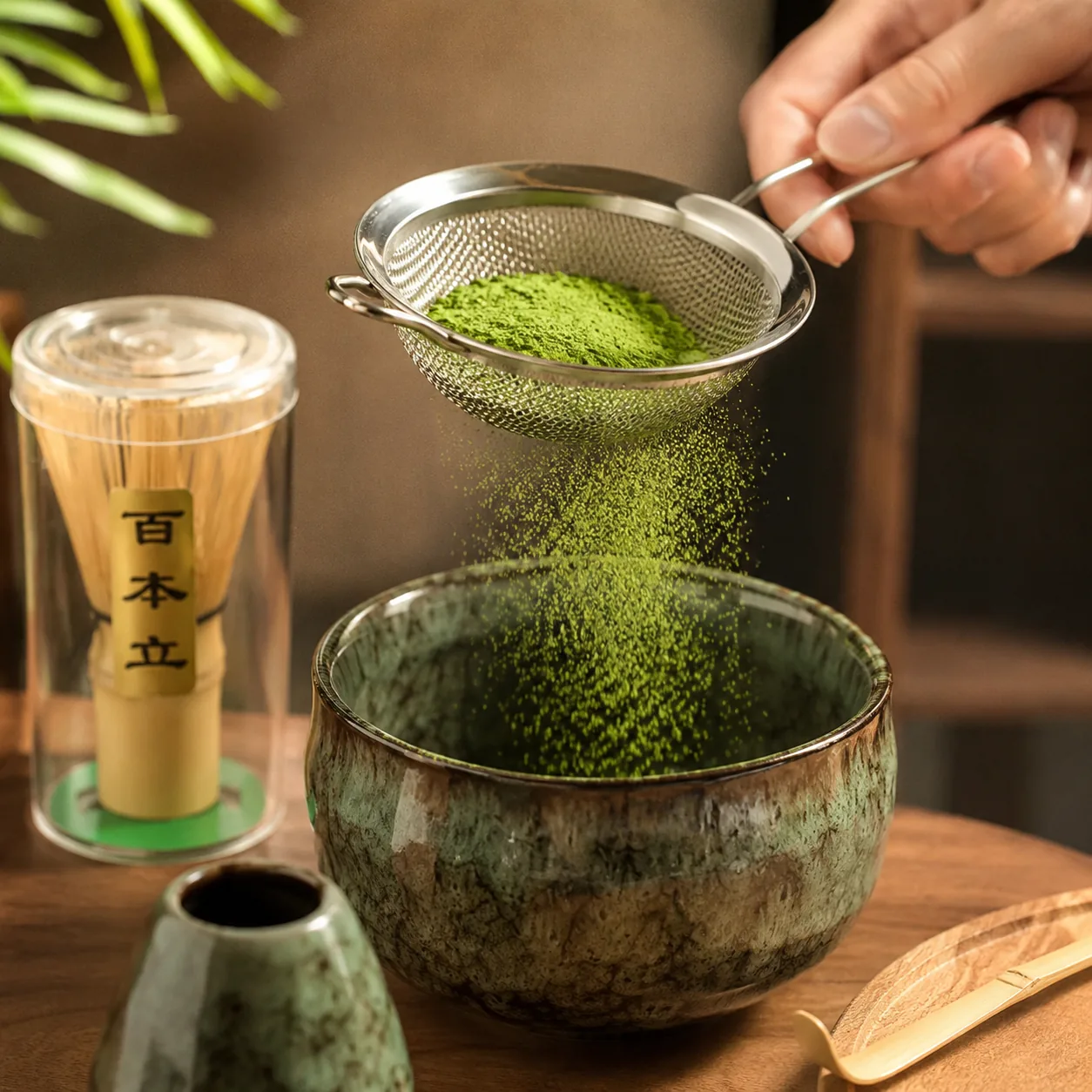 Sifting bright green matcha powder through a fine strainer into a tall iced matcha glass beside a green ceramic bowl and a bamboo whisk