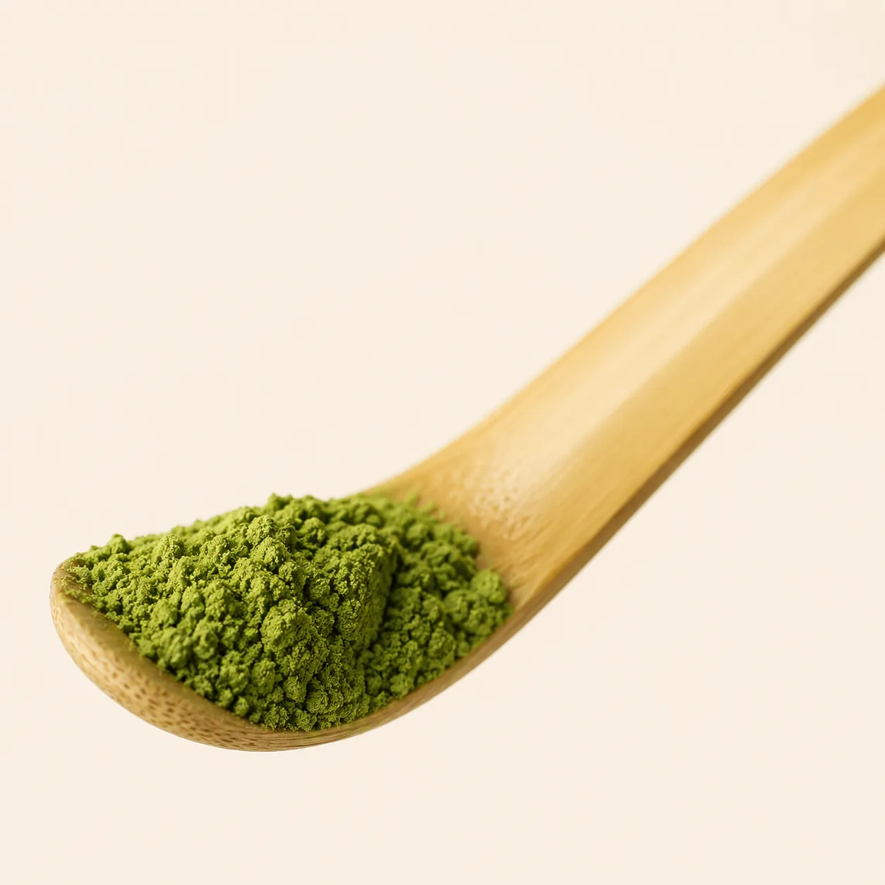 A curved bamboo chashaku scoop holding a small mound of bright green matcha powder against a cream background