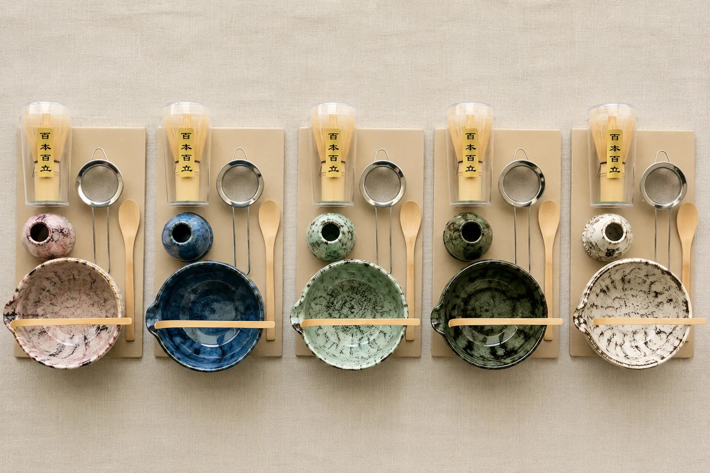 Five complete matcha sets photographed from above and arranged in rows by color on kraft boards, each laid out with its ceramic bowl, whisk holder, bamboo whisk in a tube, sifter and spoon